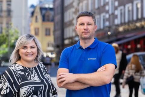 A man and a woman stand smiling in front of blurred pedestrians and buildings.