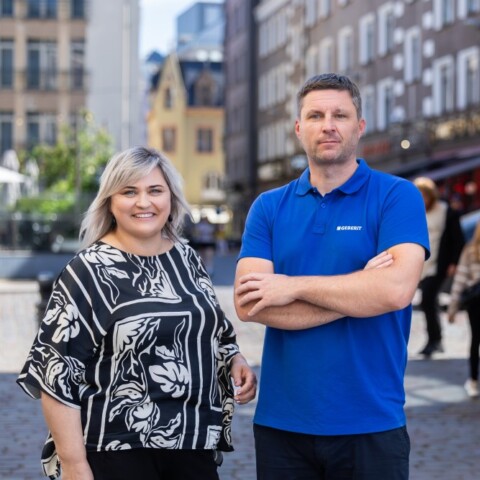 A woman in a patterned black and white blouse stands beside a man in a blue polo shirt, both smiling in a street with buildings and people in the background.