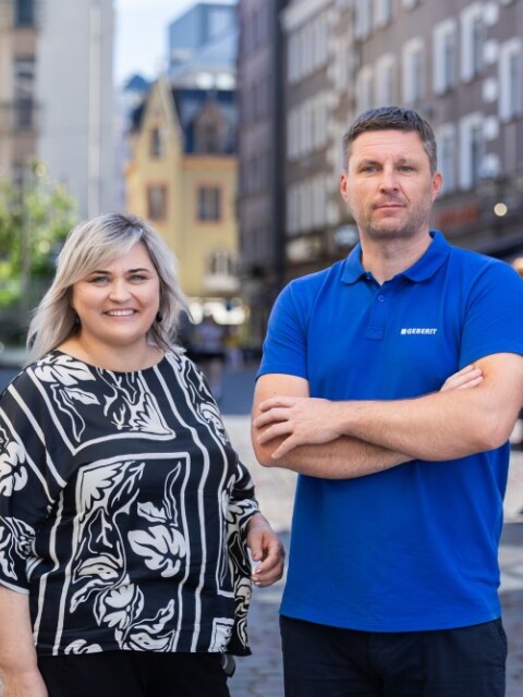 A woman in a patterned black and white blouse stands beside a man in a blue polo shirt, both smiling in a street with buildings and people in the background.