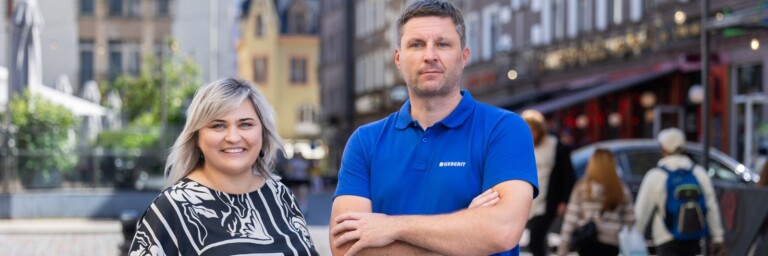 A man and a woman stand smiling in front of blurred pedestrians and buildings.