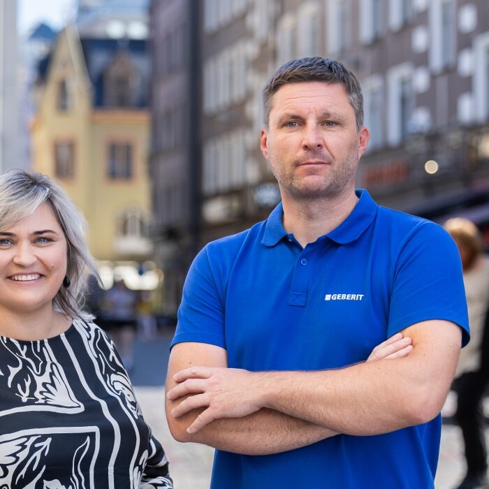 A man and a woman stand smiling in front of blurred pedestrians and buildings.