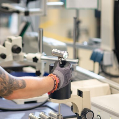 A person in a glove holding a small device while working in a laboratory setting with various scientific equipment.