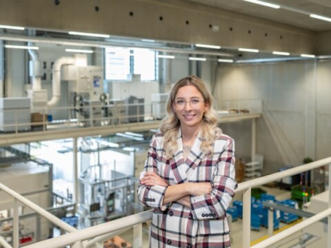 A smiling woman with wavy hair stands with arms crossed in a modern industrial setting, surrounded by machinery and equipment.