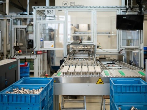 A manufacturing assembly line with conveyor belts and blue bins filled with metal parts, surrounded by machinery.