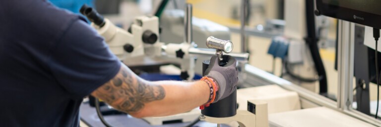 A person wearing a black shirt and gloves is operating a small device in a laboratory setting with microscopes and other equipment.