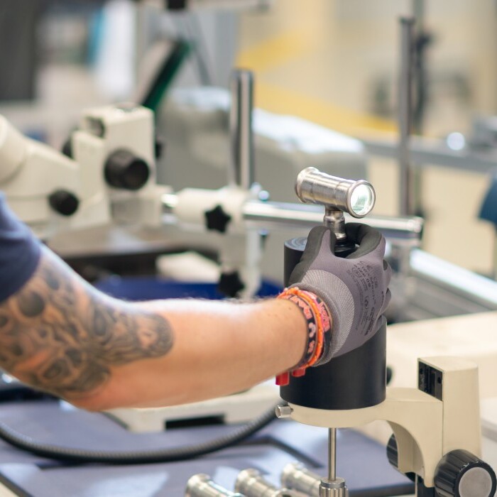 A person wearing a black shirt and gloves is operating a small device in a laboratory setting with microscopes and other equipment.