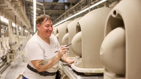 Eine Person in Arbeitskleidung bemalt keramische WC-Rohlinge in einer Fabrikhalle.