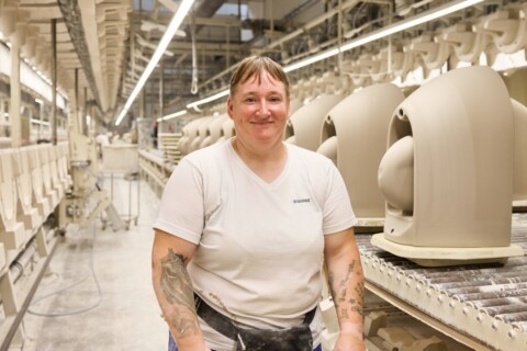 A smiling person with stands in a manufacturing facility beside rows of beige ceramics on a conveyor belt.