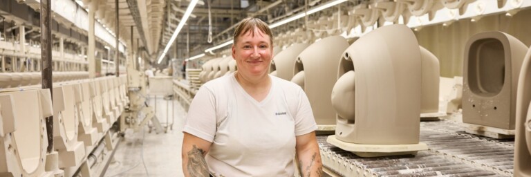 A smiling person with stands in a manufacturing facility beside rows of beige ceramics on a conveyor belt.