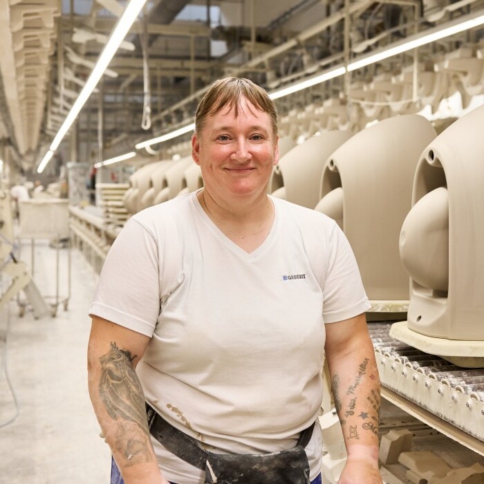 A smiling person with stands in a manufacturing facility beside rows of beige ceramics on a conveyor belt.