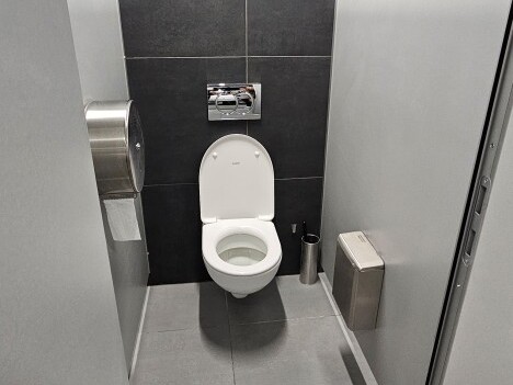 Eine Damen- oder Herrentoilette mit einem weissen Toilettenbecken, grauen Fliesen und einem Wandspender für Toilettenpapier.