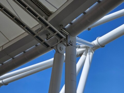 Nahaufnahme von weissen Metallstützen und Verbindungsdetails unter einem Dach, vor blauem Himmel.