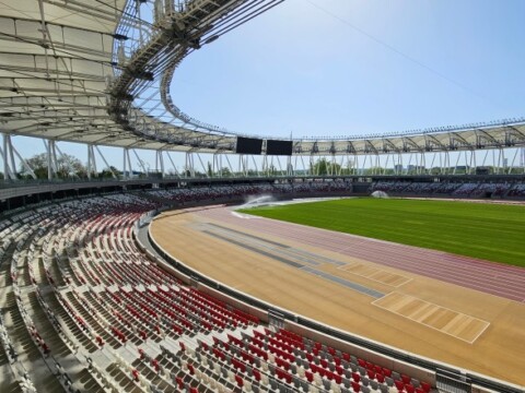 Ein modernes Sportstadion mit einer Laufbahn, grüner Rasenfläche und leeren Sitzplätzen unter klarem Himmel.