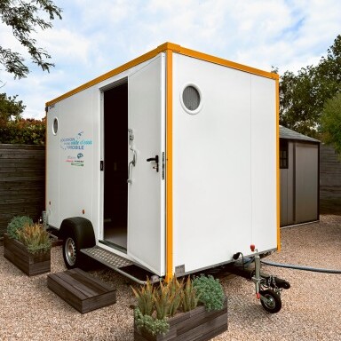 Weisser mobiler Anhänger mit gelben Akzenten, umgeben von Pflanzen und Kies, unter einem bewölkten Himmel.