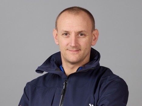 Ein Mann mit kurzem, glattem Haar trägt eine sportliche Jacke und lächelt vor einem grauen Hintergrund.