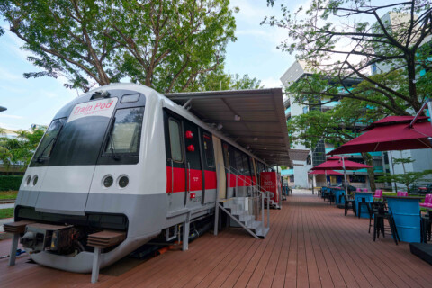 Ein stillstehender, moderner Zug mit grau-rot-weisser Lackierung, der unter einem Überstand steht, umgeben von Tischen und Stühlen auf einem Holzdeck.