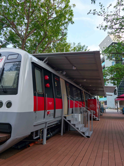 Ein stillstehender, moderner Zug mit grau-rot-weisser Lackierung, der unter einem Überstand steht, umgeben von Tischen und Stühlen auf einem Holzdeck.