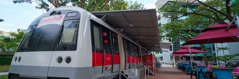 Ein stillstehender, moderner Zug mit grau-rot-weisser Lackierung, der unter einem Überstand steht, umgeben von Tischen und Stühlen auf einem Holzdeck.