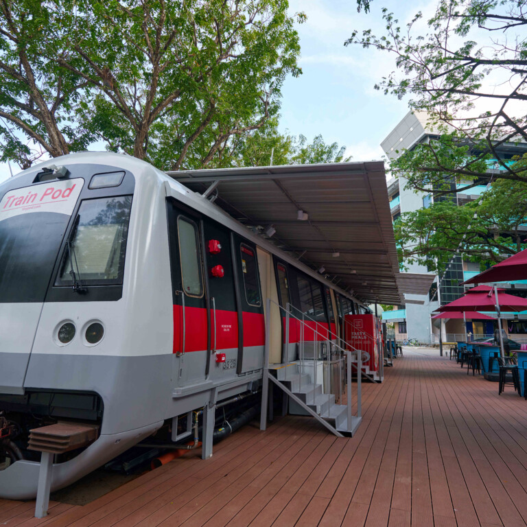 Ein stillstehender, moderner Zug mit grau-rot-weisser Lackierung, der unter einem Überstand steht, umgeben von Tischen und Stühlen auf einem Holzdeck.