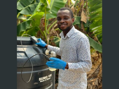 A person wearing a patterned shirt and blue gloves stands next to a large black container in a lush, green environment, holding a device.