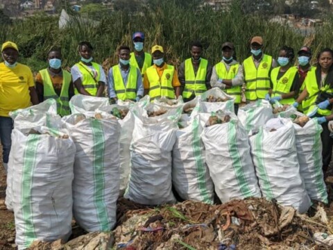 A group of people wearing safety vests and masks stands in front of large bags filled with collected waste, with a green landscape in the background.