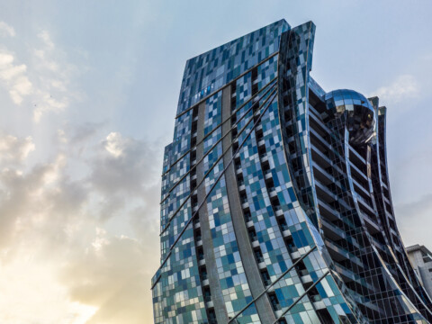 Moderne Bürogebäude mit schimmernder Glasfassade und geschwungener Architektur, umgeben von einer urbanen Landschaft und einem Himmel im Abendlicht.