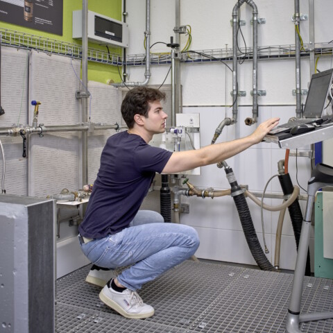 Ein junger Mann in blauer Jeans und schwarzem T-Shirt, der in einer technischen Umgebung an einem Laptop arbeitet.