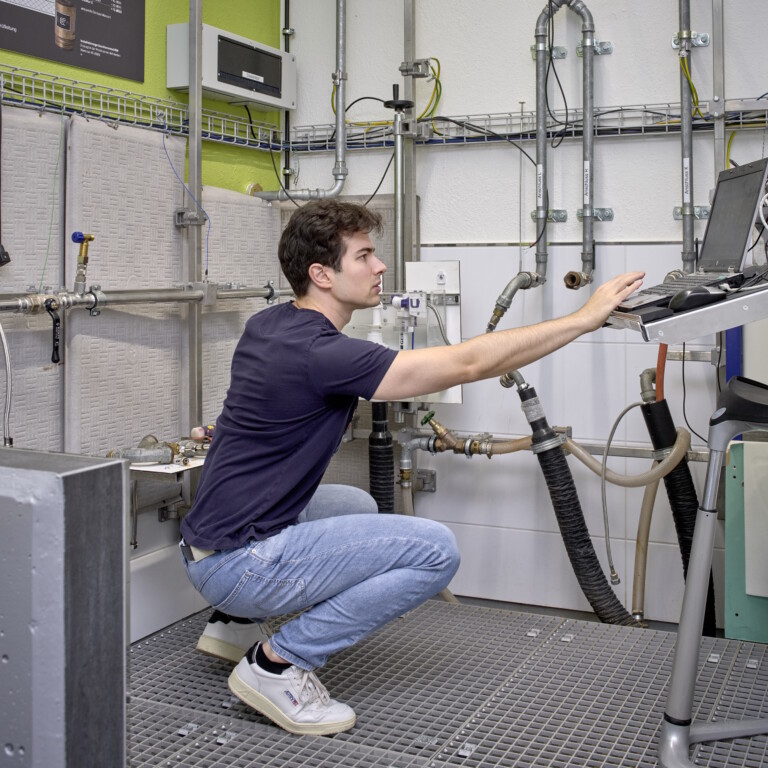 Ein junger Mann in blauer Jeans und schwarzem T-Shirt, der in einer technischen Umgebung an einem Laptop arbeitet.