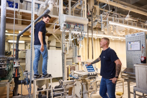 Zwei Personen in Arbeitskleidung stehen in einer industriellen Umgebung mit Maschinen und Rohrleitungen.
