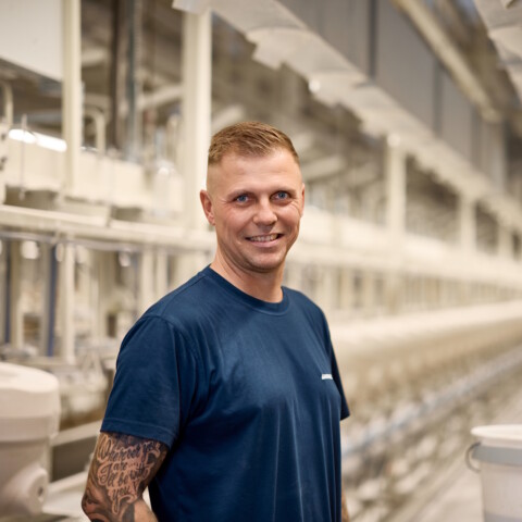 Ein lächelnder Mann steht in einer modernen Produktionshalle mit langen Maschinenreihen im Hintergrund.