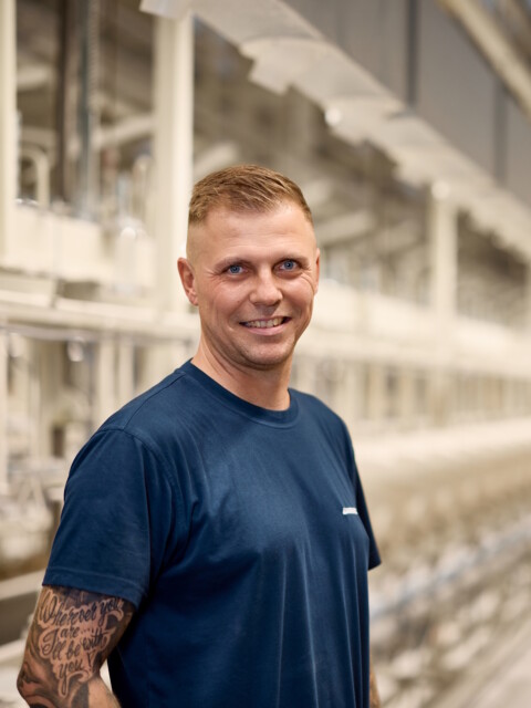 Smiling man in a blue shirt stands in a large industrial facility with long rows of machinery behind him.