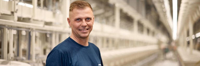 Ein lächelnder Mann steht in einer modernen Produktionshalle mit langen Maschinenreihen im Hintergrund.
