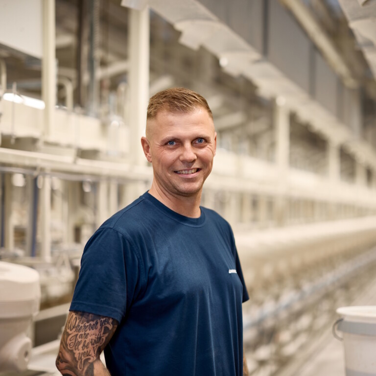 Ein lächelnder Mann steht in einer modernen Produktionshalle mit langen Maschinenreihen im Hintergrund.