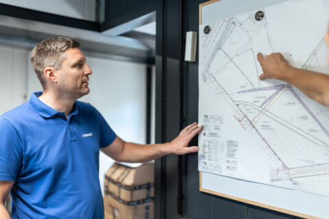 A man in a blue shirt stands next to a wall-mounted plan, pointing at details while another hand gestures towards areas of interest.