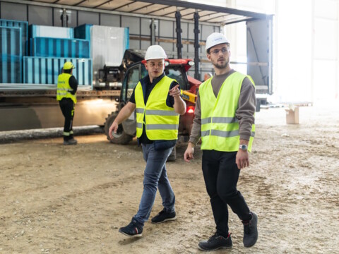 Two men wearing safety vests and hard hats walk across a construction site while discussing.