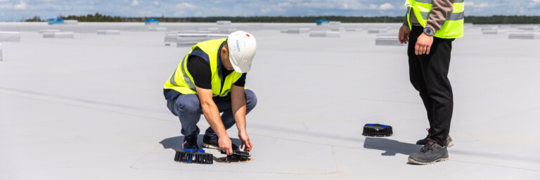 Two construction workers in safety vests and helmets, one kneeling on a flat rooftop while working with equipment, and the other standing and observing under a cloudy sky.