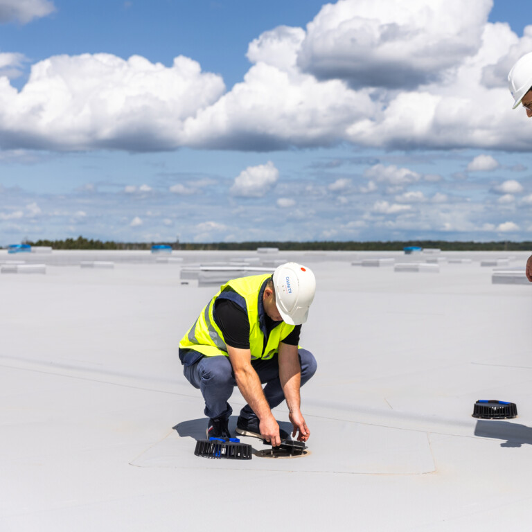 Two construction workers in safety vests and helmets, one kneeling on a flat rooftop while working with equipment, and the other standing and observing under a cloudy sky.