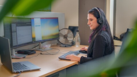A woman wearing headphones sits at a desk with multiple monitors, working on a computer in a bright office environment surrounded by greenery.