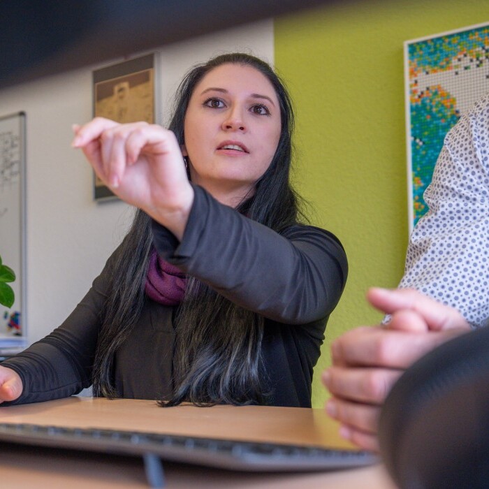 A woman with long dark hair gestures while discussing something with a man in a professional office setting.