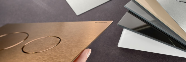 Eine Hand hält eine bronzefarbene Betätigungsplatte mit runden Tasten. Auf der rechten Seite hat es weitere Platten mit verschiedenen Materialien und Farben.