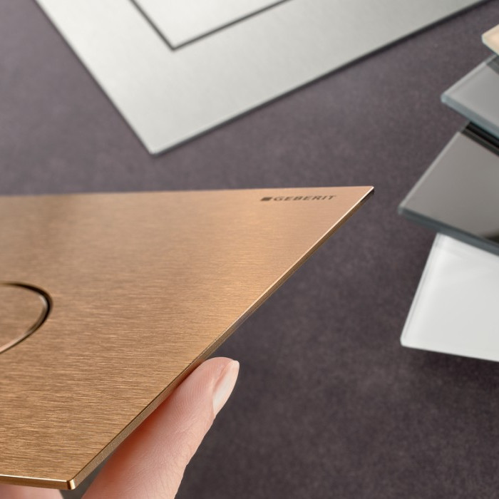 Eine Hand hält eine bronzefarbene Betätigungsplatte mit runden Tasten. Auf der rechten Seite hat es weitere Platten mit verschiedenen Materialien und Farben.