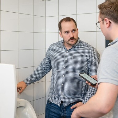 Zwei Männer stehen in einem modernen Badezimmer neben einem Urinal, einer hält ein Smartphone und schaut den anderen an.