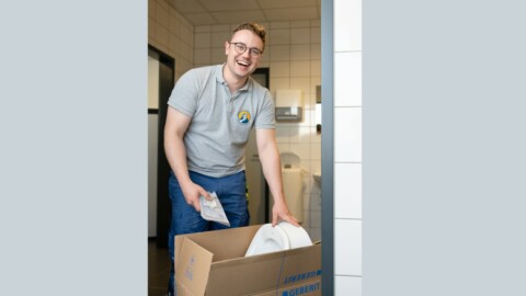 Ein junger Mann mit Brille lächelt und steht hinter einem Karton, worin ein Urinal ist.