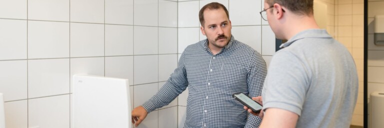 Zwei Männer in einem modernen Badezimmer, einer steht vor einem Urinal und der andere hält ein Smartphone in der Hand.