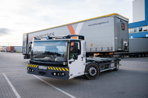 A specialized lorry with a flatbed, parked in a loading area beside a large trailer.