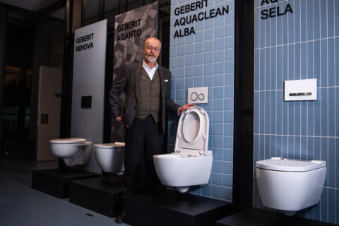 A man in a formal outfit stands next to several modern toilet models displayed in a showroom.