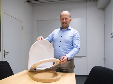 A man in a blue shirt demonstrates a modern toilet seat design while standing at a table in a minimalist office setting.