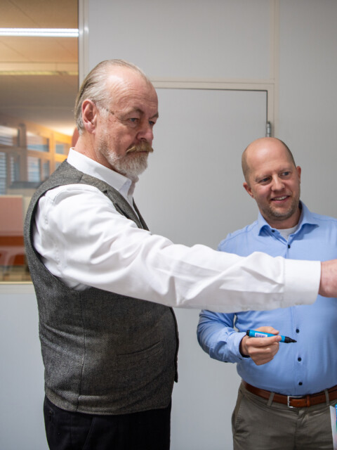 Two men discussing diagrams on a flip chart in an office setting, one pointing at the chart while the other listens attentively.
