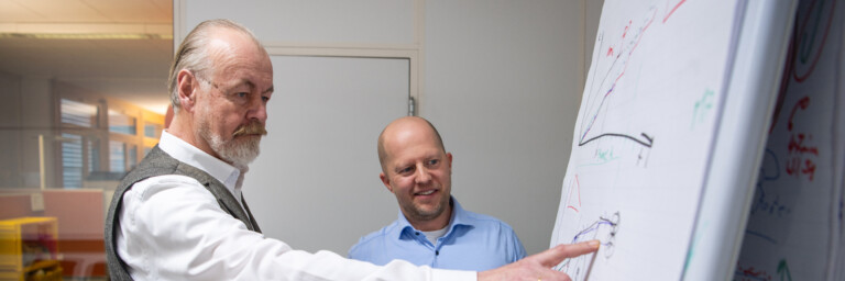 Two men discussing diagrams on a flip chart in an office setting, one pointing at the chart while the other listens attentively.