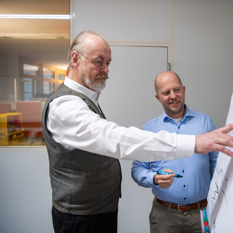 Two men discussing diagrams on a flip chart in an office setting, one pointing at the chart while the other listens attentively.